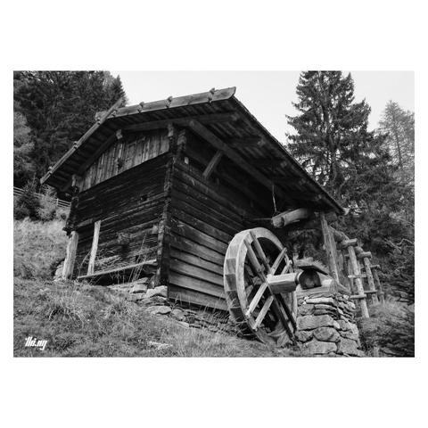 B&W photo of a ~500 years old wooden, watermill on a steep hillside. The big water wheel is mounted on the outside of the house and the supply pipe (also wooden) is hanging from the roof. Forest in the background.